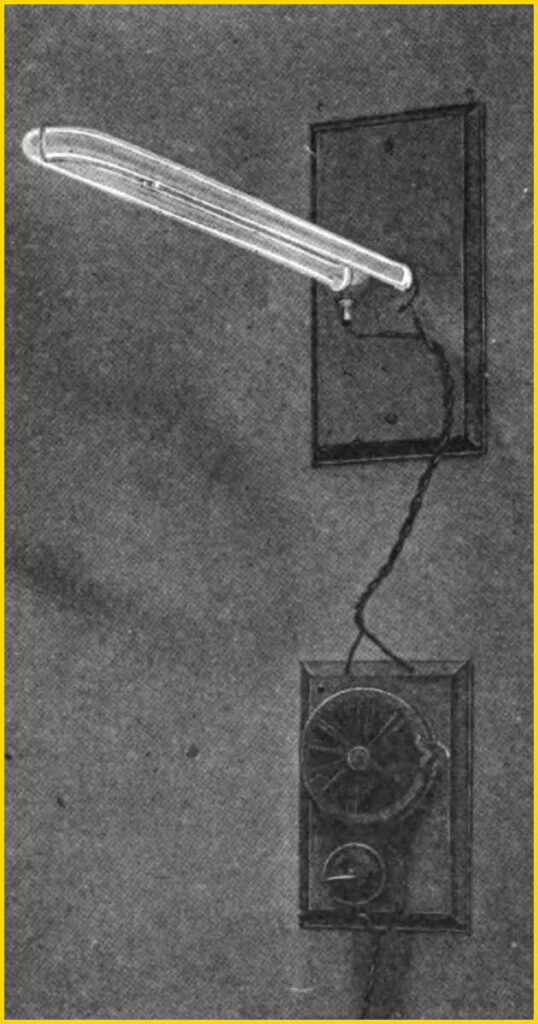 A historic photograph of Peter Cooper Hewitt's first mercury vapor lamp from 1903. The image shows a long, horizontal glass tube with electrodes, positioned above a circular electrical ballast. This invention was the direct predecessor of the modern fluorescent light.